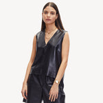 Eine Frau in einem ärmellosen schwarzen Ledertop und einer Hose, die mit Goldschmuck wie der LIEBESKIND BERLIN Halskette - The Nugget von Liebeskind Berlin verziert ist, steht vor einem weißen Hintergrund und verkörpert mühelosen urbanen Stil.