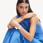 Eine Frau in einem blauen Outfit sitzt selbstbewusst mit verschränkten Armen da, trägt eine Uhr, bunte Armbänder und stilvolle Liebeskind Berlin Creole - Keep Loving Ohrringe und schaut direkt in die Kamera.