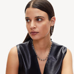 A woman with straight, dark hair wears a sleeveless black leather top and silver earrings, accessorized with the LIEBESKIND BERLIN Halskette – The Knot. She gazes to the side with a neutral expression against a plain background.