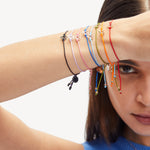 Eine Frau legt bunte Kettenarmbänder mit geometrischen Anhängern und ein LIEBESKIND BERLIN Armband - Keep Loving übereinander und verdeckt damit teilweise ihr Gesicht für einen schicken Accessoire-Look.