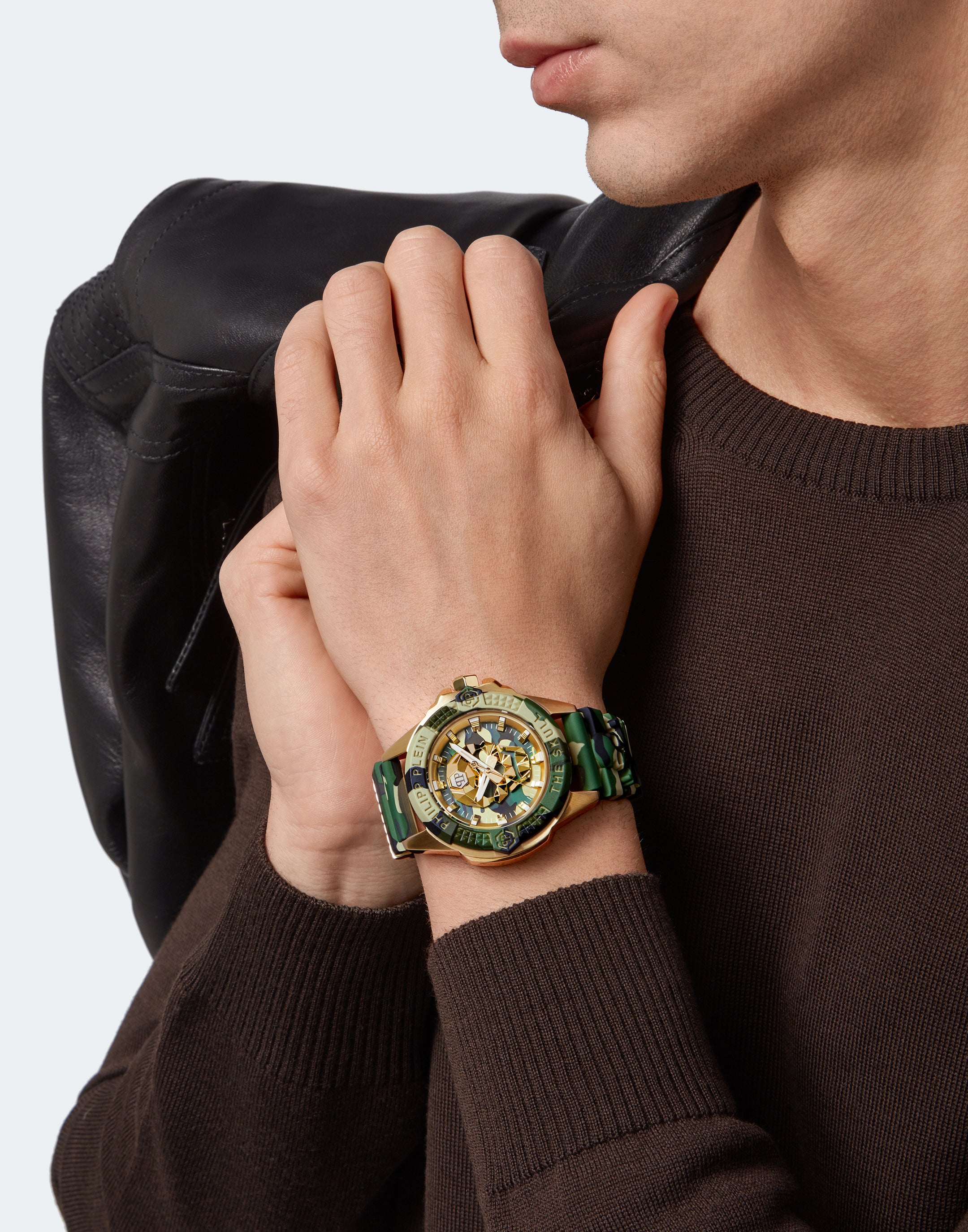 Mann in einem braunen Pullover, der die Philipp Plein Armbanduhr - The Golden Camouflage trägt und eine schwarze Jacke über der Schulter hält.