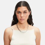 Frau mit langen dunklen Haaren in einem weißen Tank-Top, kombiniert mit der Liebeskind Berlin Halskette - Big Link und passenden silbernen Ohrringen.