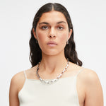 Frau mit langen gewellten Haaren, die eine klobige Liebeskind Berlin Big Link-Halskette und ein weißes ärmelloses Oberteil trägt.