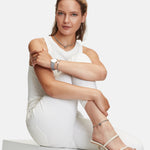 Eine Frau in einem weißen Outfit posiert sitzend vor einem schlichten Hintergrund, trägt Sandalen und Schmuck, um den Ear Candy Look mit dem Broken Heart Mini Ohrstecker von Liebeskind Berlin zu präsentieren.