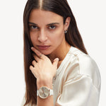 Eine Frau in einer cremefarbenen Bluse trägt Liebeskind Berlin Ohrstecker - The Big Knot Ohrringe, eine weiße Armbanduhr und einen Ring und schaut direkt in die Kamera.