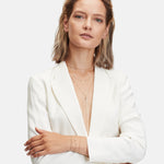 Eine Frau in einem weißen Anzug und mit mehreren Ketten trägt die Ohrringe LIEBESKIND BERLIN Creole - Gold Collection von Liebeskind Berlin für einen Ear Candy Look. Sie hat hellbraunes Haar und steht mit verschränkten Armen vor einem schlichten Hintergrund.