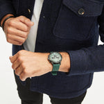 Eine Person in einem marineblauen Mantel trägt eine Fynch-Hatton-Armbanduhr - The Green Chrono an ihrem linken Handgelenk vor einem weißen Hintergrund.