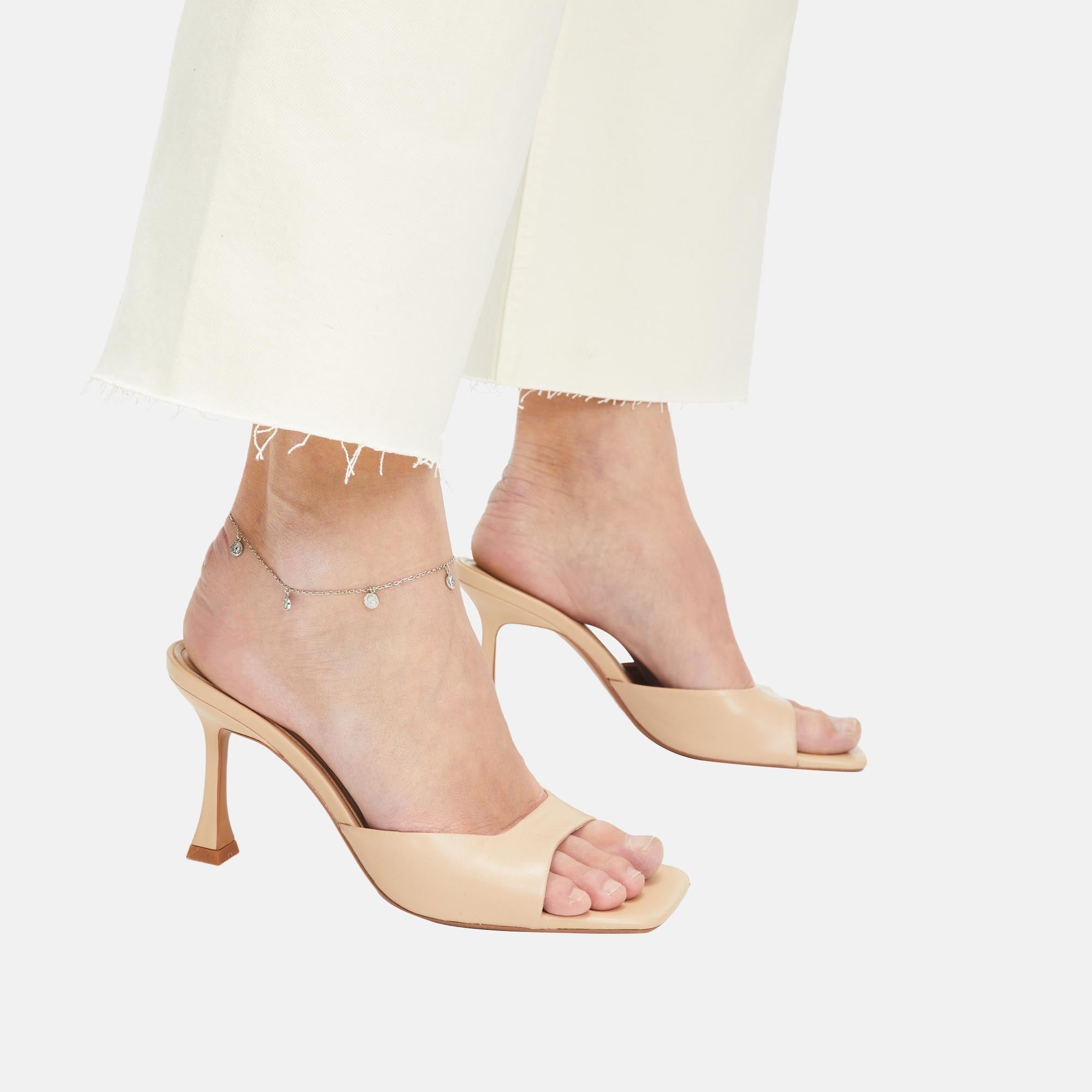 Eine Frau trägt cremefarbene Tamaris-Sandalen mit Absatz und eine weiße Hose mit ausgefranstem Saum, die mit der Tamaris Fußkette - Coin by Tamaris an einem Knöchel verziert ist.
