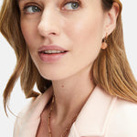 Frau mit Tamaris Creole - Coin rosegoldene Ohrringe und Halskette von Tamaris, bekleidet mit einem blassrosa Blazer, schaut leicht zur Seite.