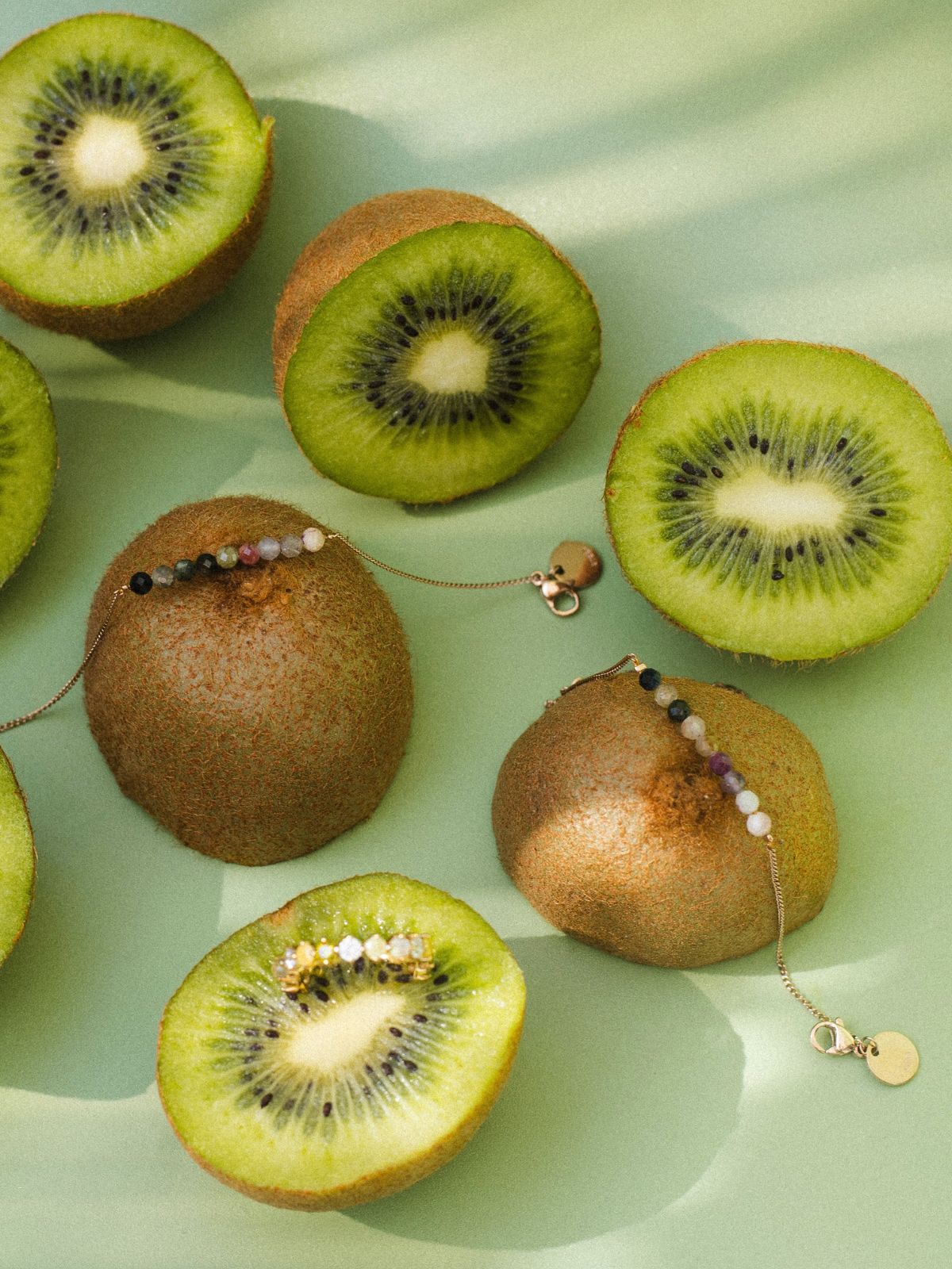 Filigrane Armbänder mit bunten Beads und Natursteinen zwischen aufgeschnittenen Kiwis auf grünem Hintergrund.