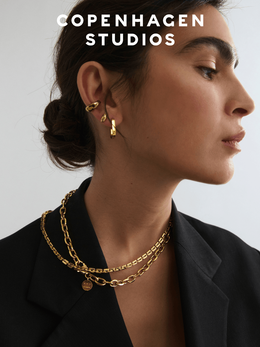  zur ersten Schmuck- und Uhrenkollektion von COPENHAGEN STUDIOS – stilvolles Portrait einer Frau mit goldenen Statement-Ohrringen und Layering-Ketten, kombiniert mit einem eleganten schwarzen Blazer.