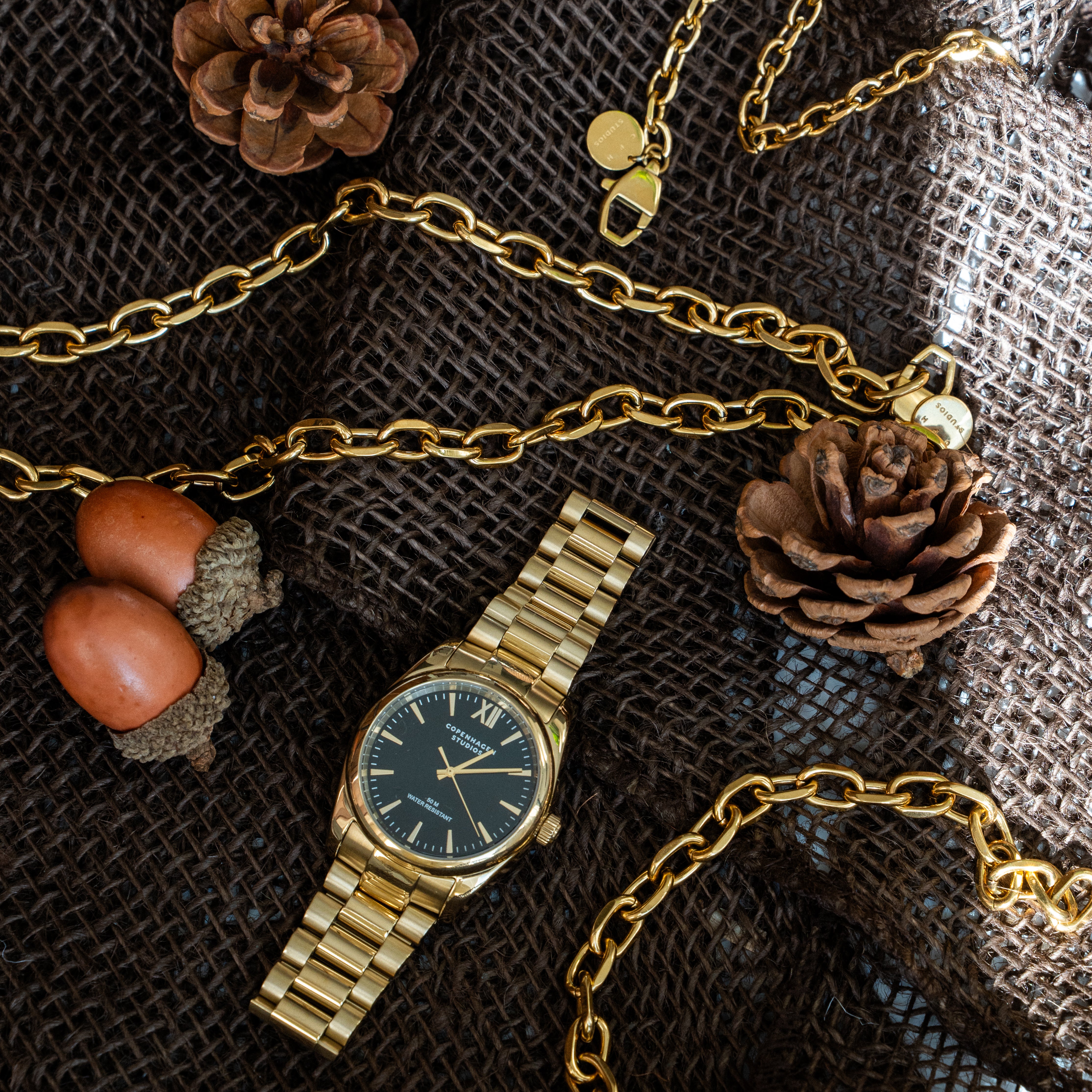 Goldfarbene COPENHAGEN STUDIOS Armbanduhr mit rundem Gehäuse, dunklem Zifferblatt und Edelstahlarmband, kombiniert mit markanten goldfarbenen Gliederketten und Armbändern. Herbstliche Schmuckinszenierung mit Tannenzapfen, Eicheln und rustikalem Jutestoff.