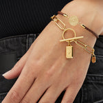 Eine mit goldenen und braunen Kettenarmbändern geschmückte Hand, darunter das Fynch-Hatton Armband - The Compass Coin von Fynch-Hatton, ruht auf einer dunklen Jeans.