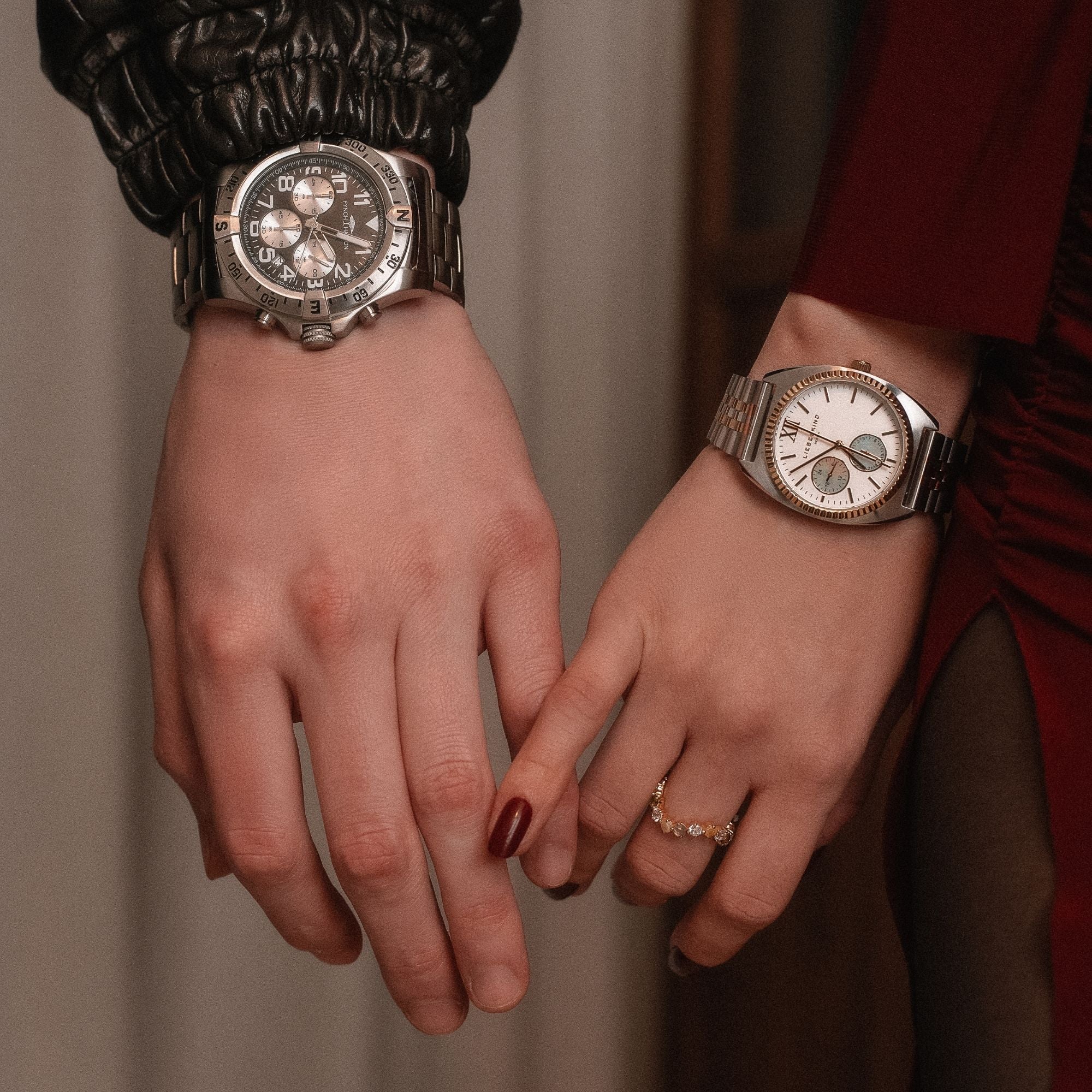 Zwei Hände, die sich berühren – eine trägt eine markante, sportliche Herrenuhr aus Edelstahl, die andere eine elegante Uhr mit hellem Zifferblatt und einem goldenen Ring. Stilvolle Geschenkidee für Männer.
