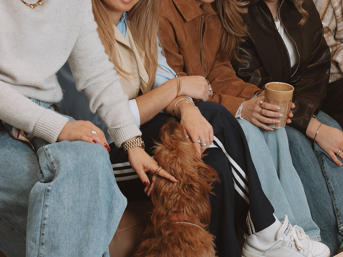Kolleginnen mit Schmuck und Uhren sitzen zusammen auf Sofa und streicheln Hund – gemütlicher Lifestyle-Moment.