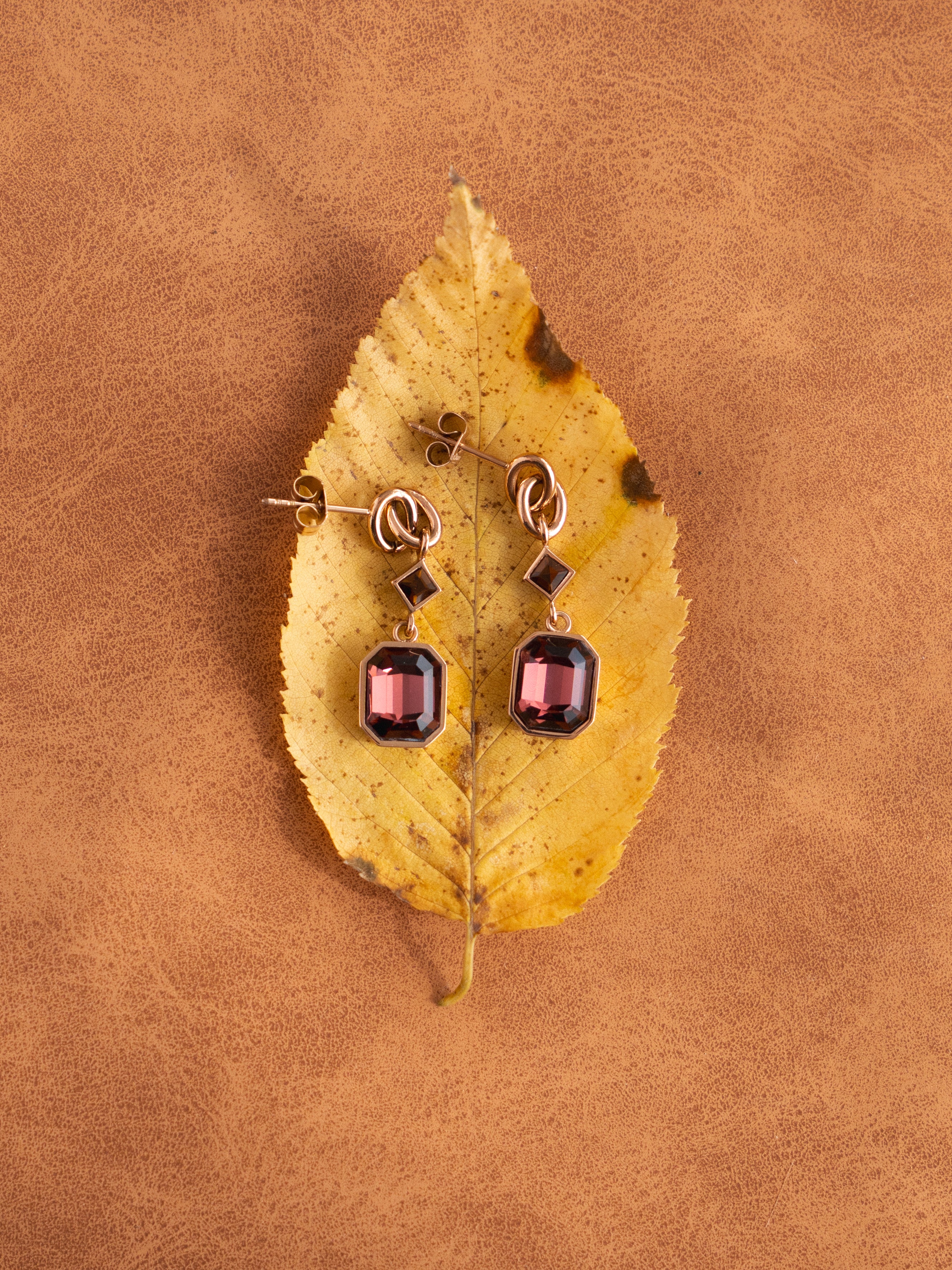 Roségoldfarbene s.Oliver Damenohrringe mit geometrischen Anhängern und facettierten, dunkelroten Schmucksteinen in rechteckiger Form. Die eleganten Ohrhänger liegen auf einem gelben Herbstblatt vor warmem, cognacbraunem Hintergrund.