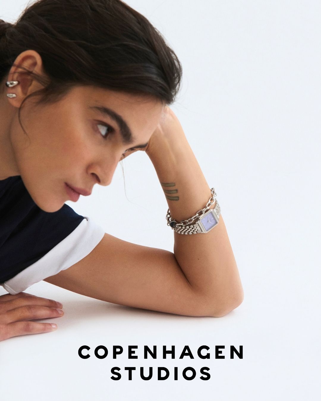 Frau mit dunklen Haaren stützt sich auf den Arm, zeigt eine Uhr und Tätowierungen, darunter der Text Copenhagen Studios.