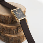 Rechteckige Uhr mit schwarzem Ziffernblatt und dem dazu passenden Silikonarmband und goldfarbenem Gehäuse von Tamaris liegt dekorativ auf Baumscheiben.