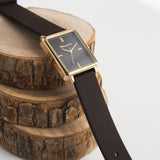 Rechteckige Uhr mit schwarzem Ziffernblatt und dem dazu passenden Silikonarmband und goldfarbenem Gehäuse von Tamaris liegt dekorativ auf Baumscheiben.