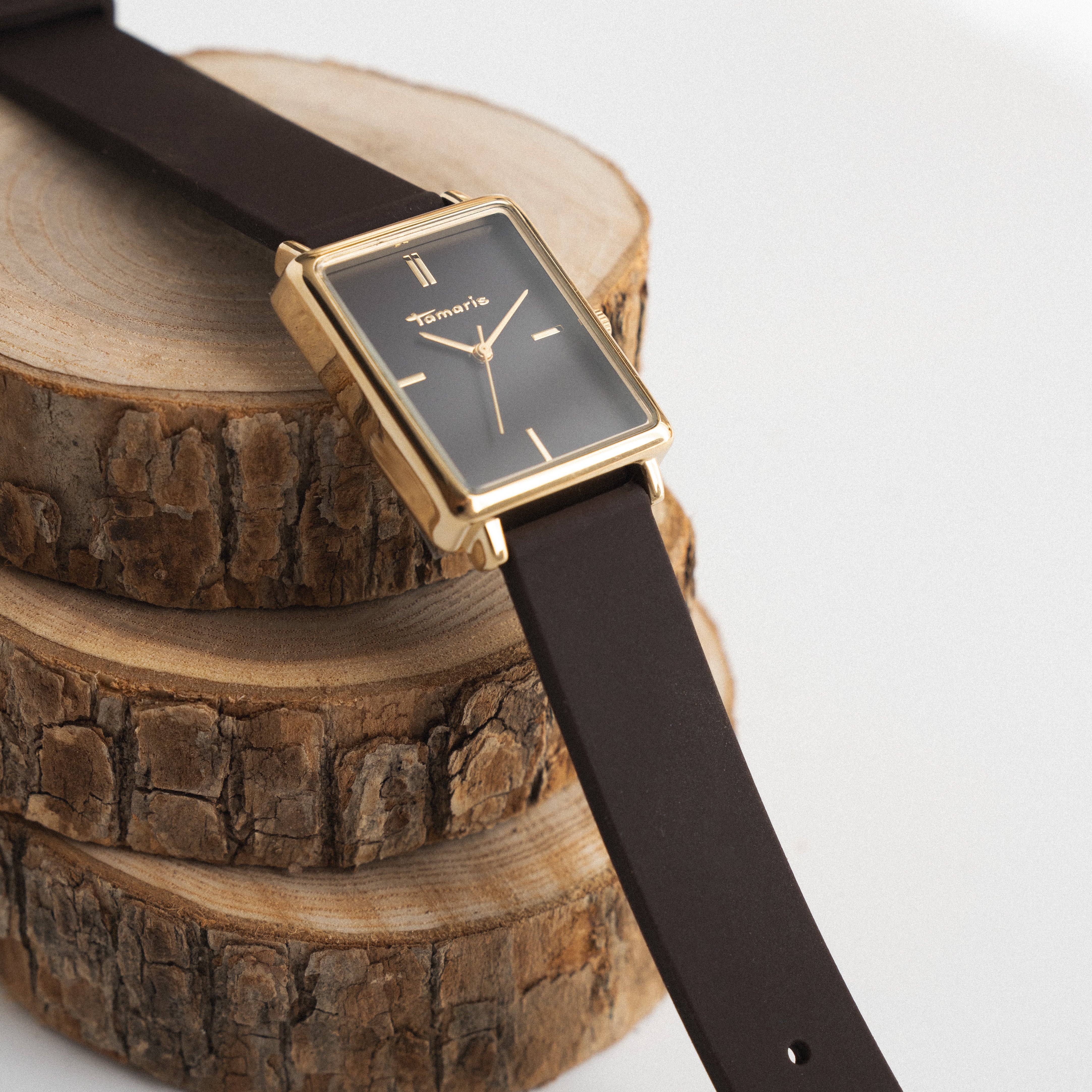 Rechteckige Uhr mit schwarzem Ziffernblatt und dem dazu passenden Silikonarmband und goldfarbenem Gehäuse von Tamaris liegt dekorativ auf Baumscheiben.