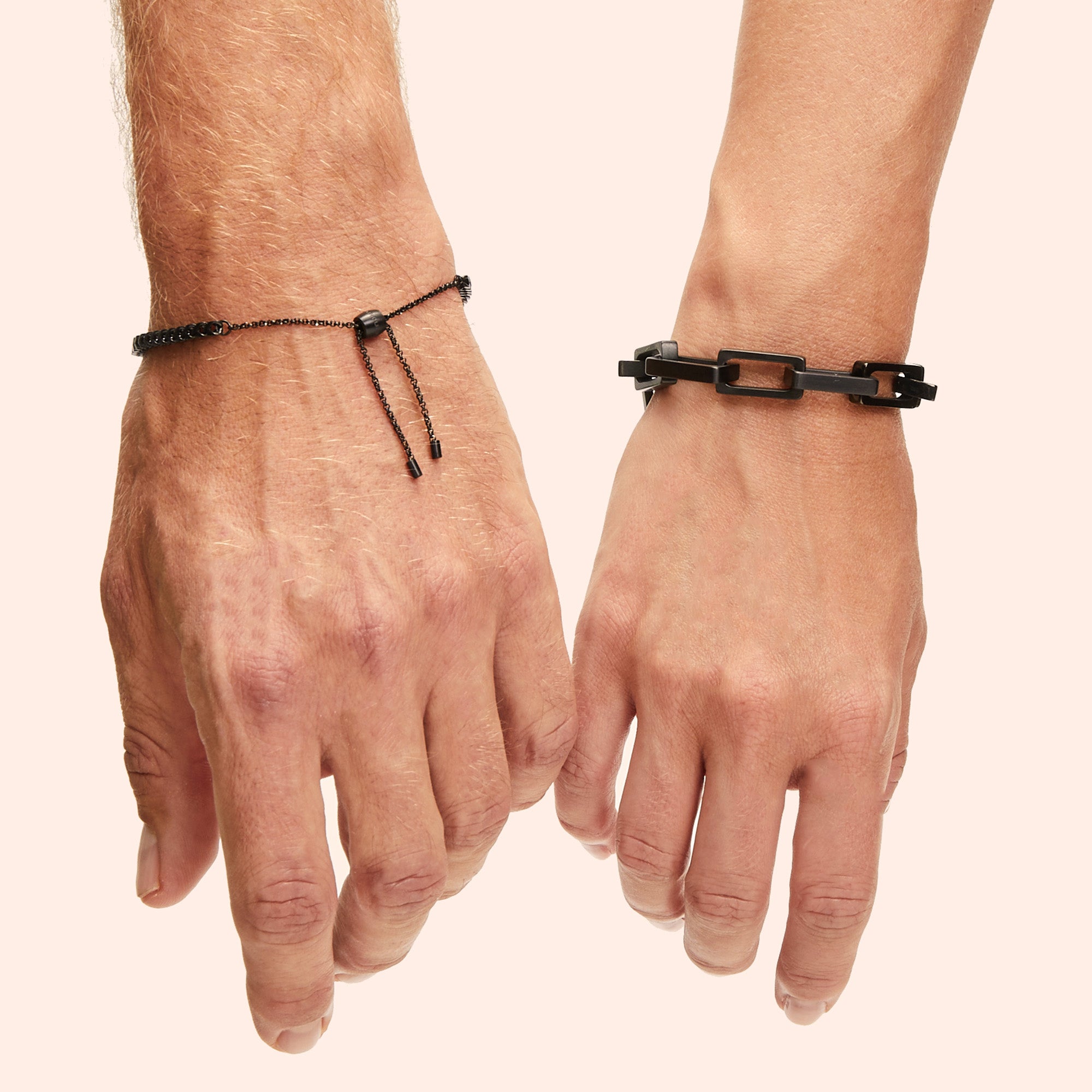 Zwei Hände mit verschiedenen schwarzen Armbändern vor einem hellbeigen Hintergrund.