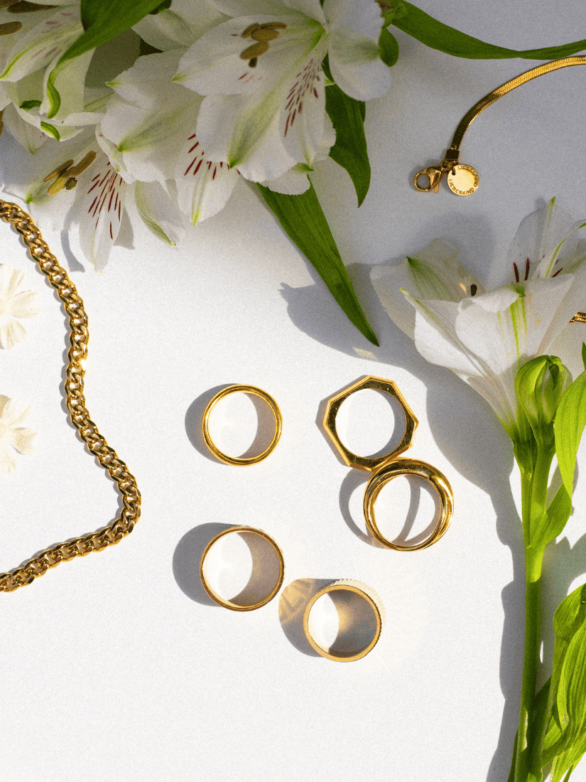 Goldfarbener Schmuck von Tamaris und Liebeskind Berlin, arrangiert neben weißen Blumen auf hellem Untergrund.