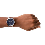 Eine Hand, die die Armani Exchange Armbanduhr - The Brown Leather mit blauem Zifferblatt, silbernem Gehäuse und braunem Lederarmband von Armani Exchange trägt, isoliert auf weißem Hintergrund.