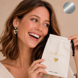 Lächelndes Model mit goldfarbenen Herzohrringen hält einen cremefarbenen cool time Schmuckbeutel mit „With Love“-Aufschrift. Die silberne Mystery Schmuckbox enthält drei Überraschungs-Schmuckstücke bekannter Marken im Wert von mindestens 165 €.