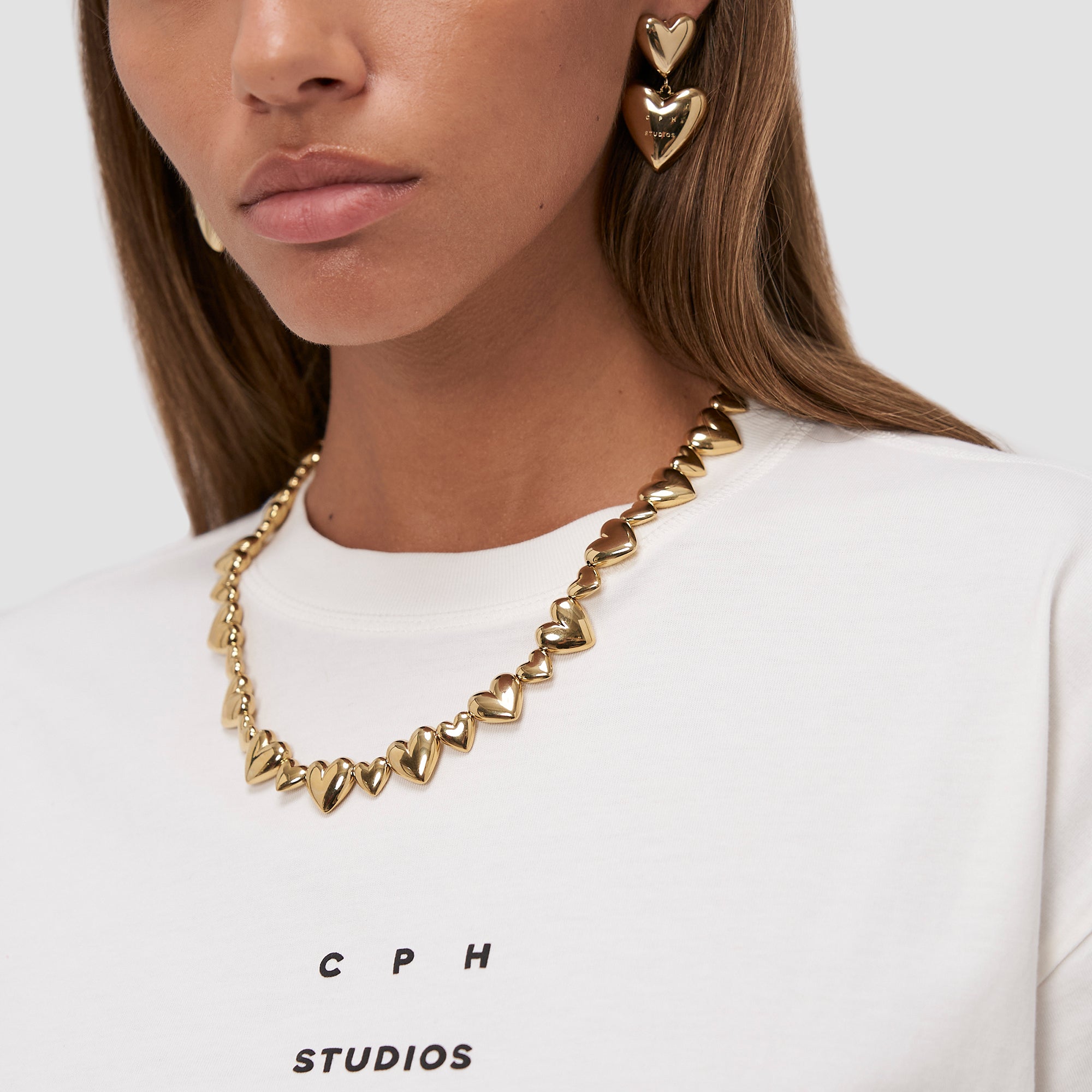 Frau trägt einen goldenen COPENHAGEN STUDIOS Puffy Hearts Ohrstecker und Halskette, gestylt mit einem weißen "COPENHAGEN STUDIOS" Hemd. Vor einem neutralen Hintergrund passt dieser elegante Ohrschmuck von Copenhagen Studios zu jedem Anlass.