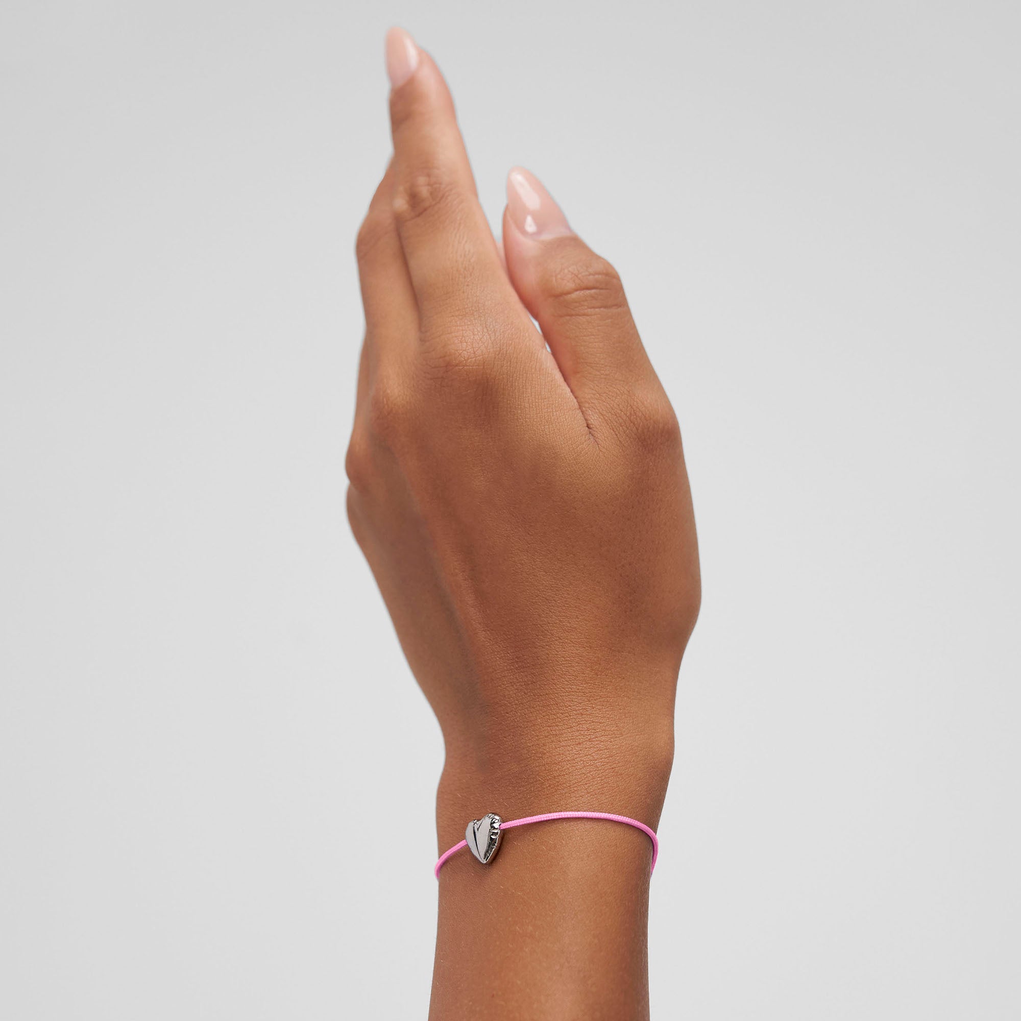 Eine Hand trägt das COPENHAGEN STUDIOS Armband - Balloon Hearts von Copenhagen Studios mit einem rosafarbenen Band und einem zarten silbernen Herzanhänger, der vor einem schlichten Hintergrund gezeigt wird.