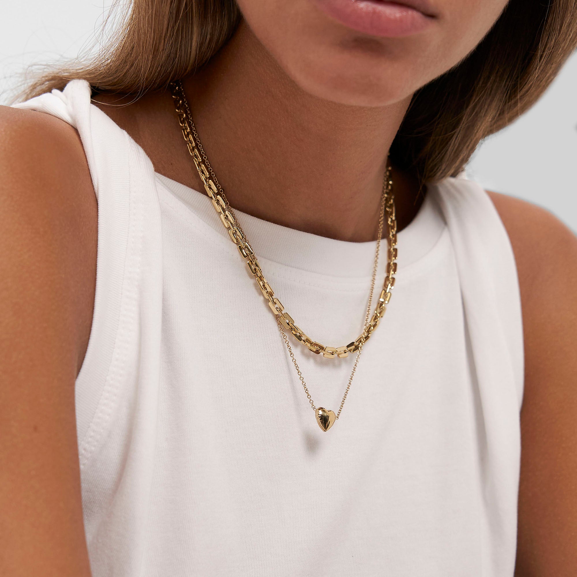 Eine Frau in einem weißen, ärmellosen Oberteil trägt zwei goldene Halsketten in Lagen - eine mit einem Herzanhänger -, die für urbane Eleganz und den Stil Edgy Necklace von Copenhagen Studios stehen und die Raffinesse der COPENHAGEN STUDIOS Halskette widerspiegeln.