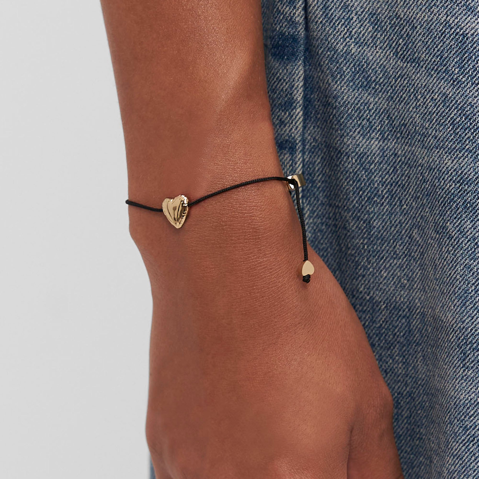 Eine Hand trägt das Copenhagen Studios Balloon Hearts Armband mit schwarzer Kordel und goldenem Herzanhänger zu einer Blue Denim Jeans.