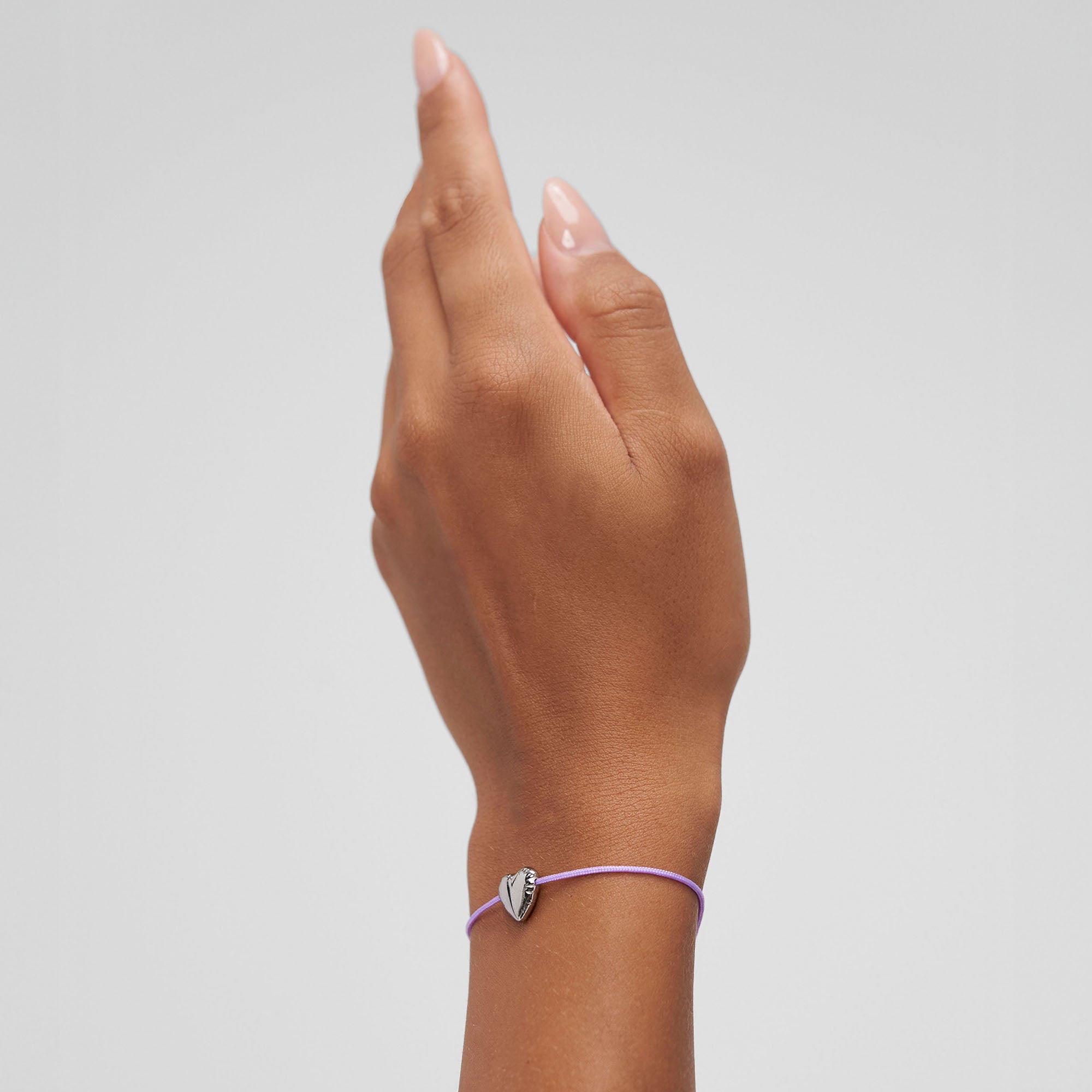 Eine Hand, die das Copenhagen Studios COPENHAGEN STUDIOS Armband - Balloon Hearts trägt. Es besteht aus einer lilafarbenen Schnur mit einem kleinen silbernen herzförmigen Anhänger.