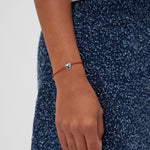 Eine Hand zeigt das COPENHAGEN STUDIOS Armband - Balloon Hearts von Copenhagen Studios mit einer roten Schnur und einem silbernen Herzanhänger, der auf einem blauen, strukturierten Rock einen eleganten Akzent setzt.