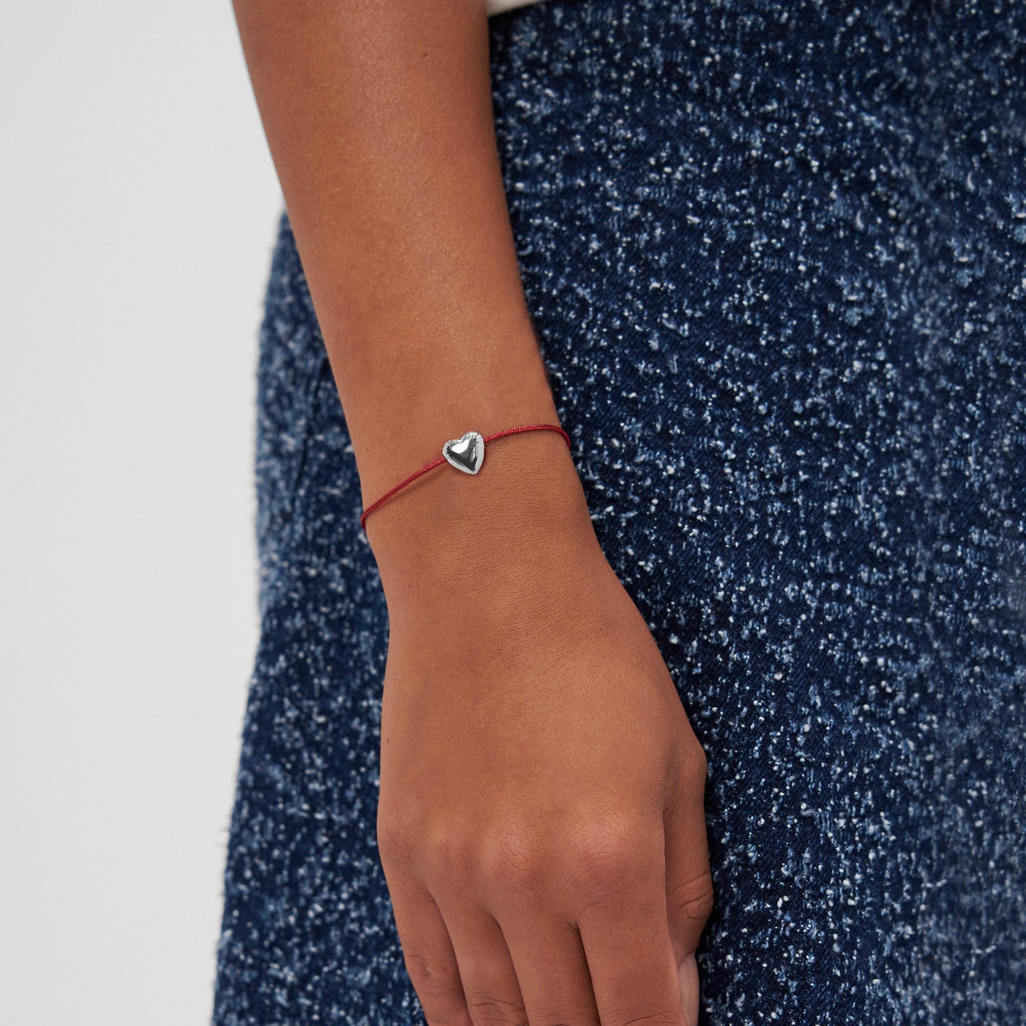 Eine Hand zeigt das COPENHAGEN STUDIOS Armband - Balloon Hearts von Copenhagen Studios mit einer roten Schnur und einem silbernen Herzanhänger, der auf einem blauen, strukturierten Rock einen eleganten Akzent setzt.