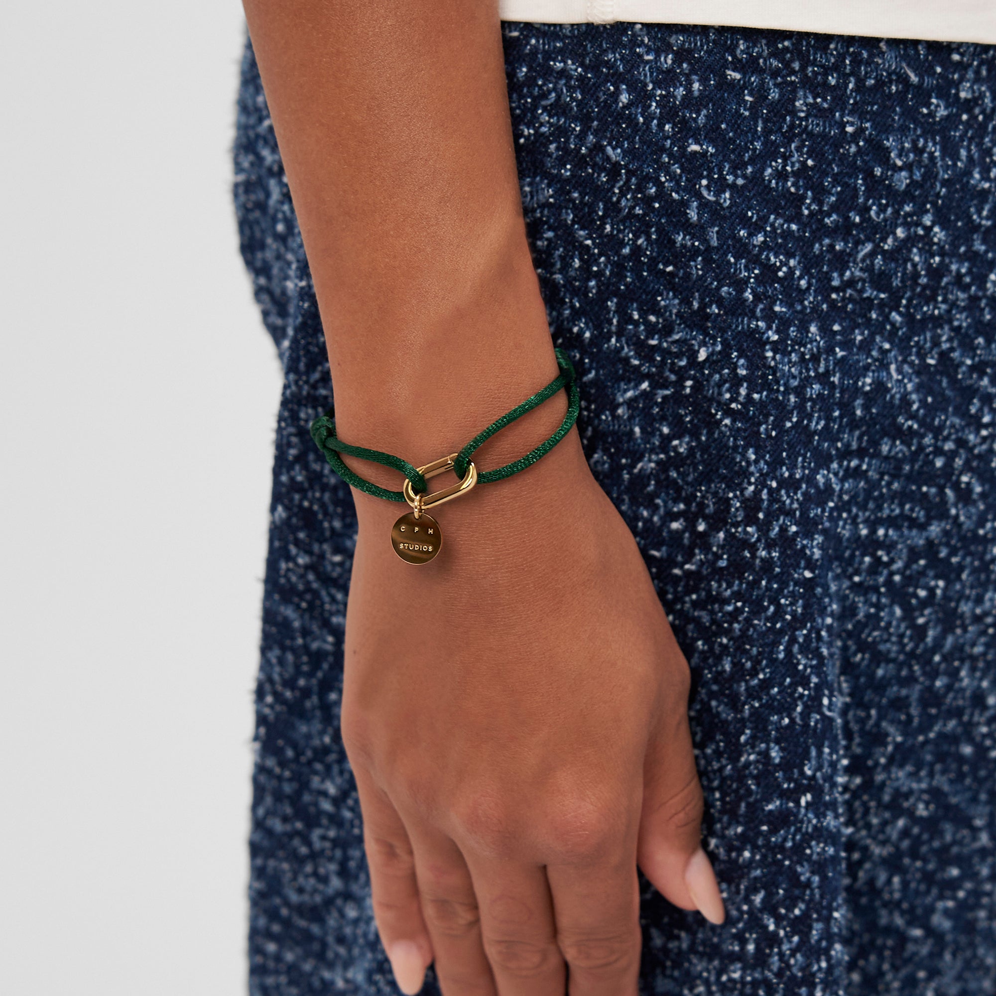 Eine Hand trägt das COPENHAGEN STUDIOS Armband - Fabric Link von Copenhagen Studios. Es besteht aus einem grünen Gliederarmband aus Stoff mit einem goldenen, kreisförmigen Anhänger, der auf blauem, strukturiertem Stoff für einen lässigen, eleganten Look sorgt.