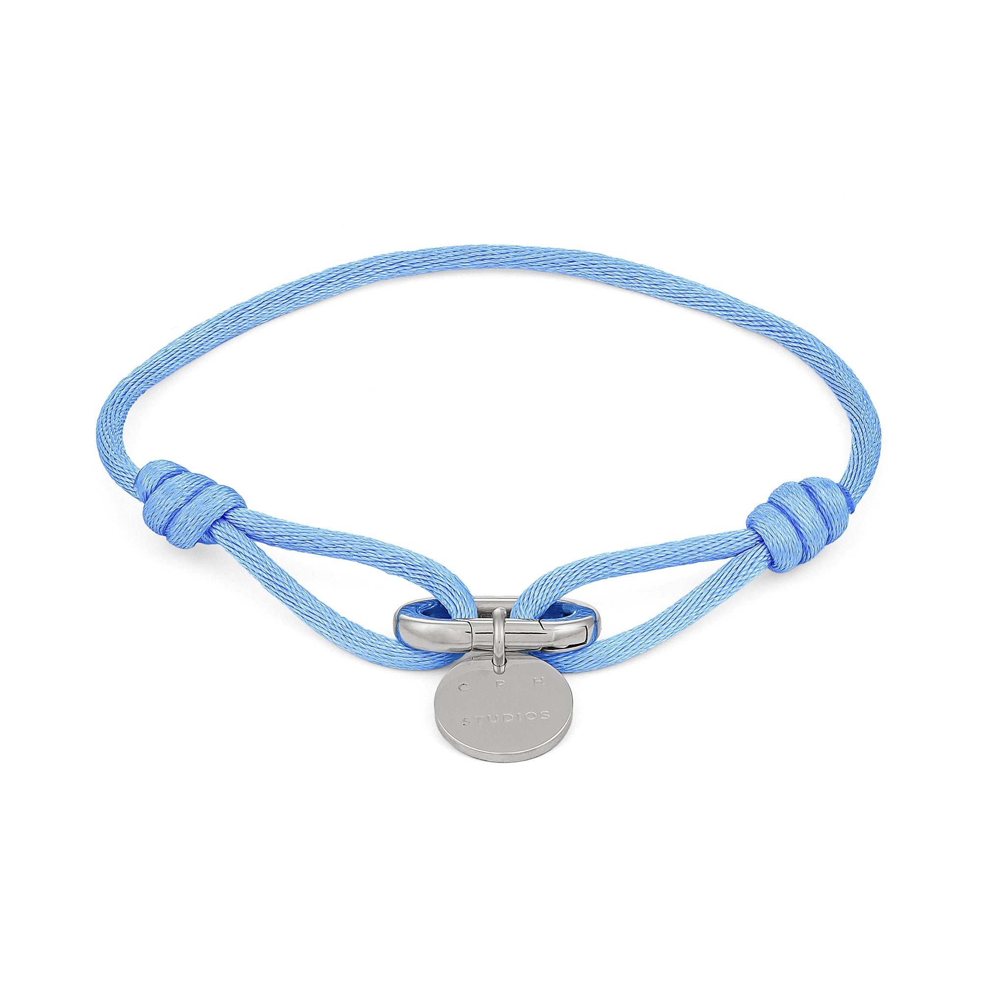 Das COPENHAGEN STUDIOS Armband - Fabric Link von Copenhagen Studios besteht aus einem hellblauen Stoffarmband mit einem runden silbernen Anhänger und einem Metallverschluss in der Mitte.