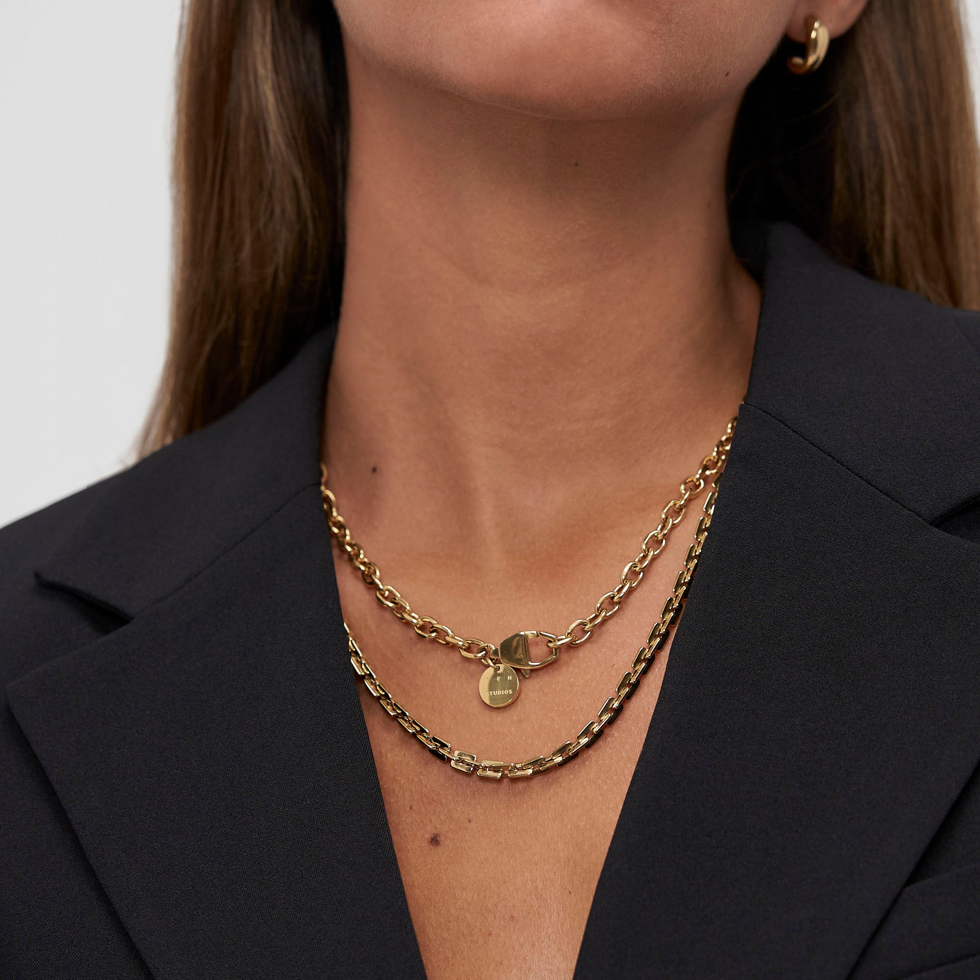 Eine Person in einem schwarzen Blazer trägt zwei übereinanderliegende Goldketten, darunter die COPENHAGEN STUDIOS Halskette - Edgy Necklace by Copenhagen Studios, sowie goldene Creolen - ein mühelos urbaner, von der Ästhetik inspirierter Look.