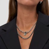 Eine Frau in einem schwarzen Blazer trägt mehrlagige Silberketten, darunter die COPENHAGEN STUDIOS Halskette - Basic Chains von Copenhagen Studios, sowie eine klobige Halskette und einen kleinen Creolenring.