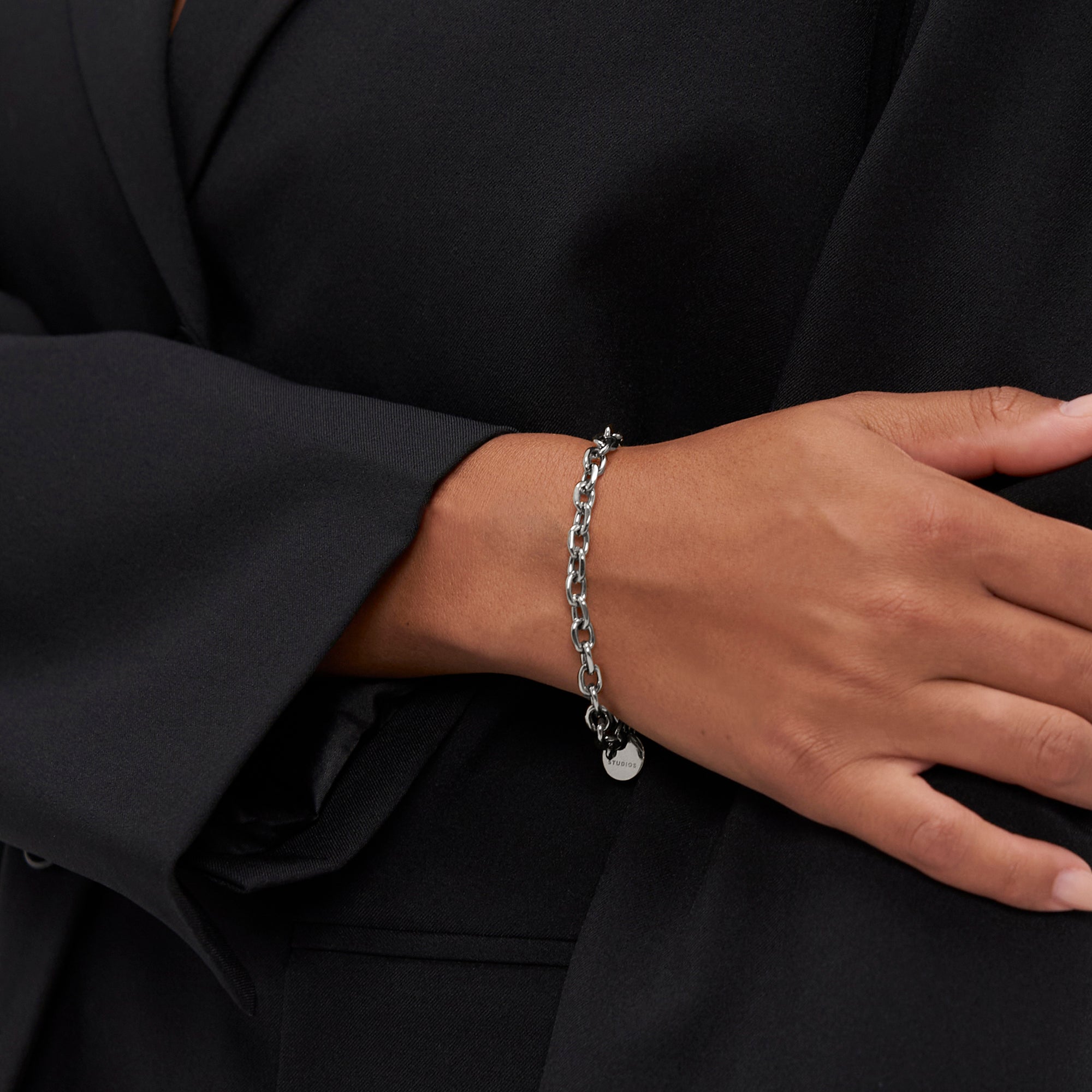 Eine Person trägt das COPENHAGEN STUDIOS Armband - Basic Chains von Copenhagen Studios zu einer schwarzen Anzugsjacke und legt eine Hand auf den Arm für einen echten Statement-Schmuck-Look.