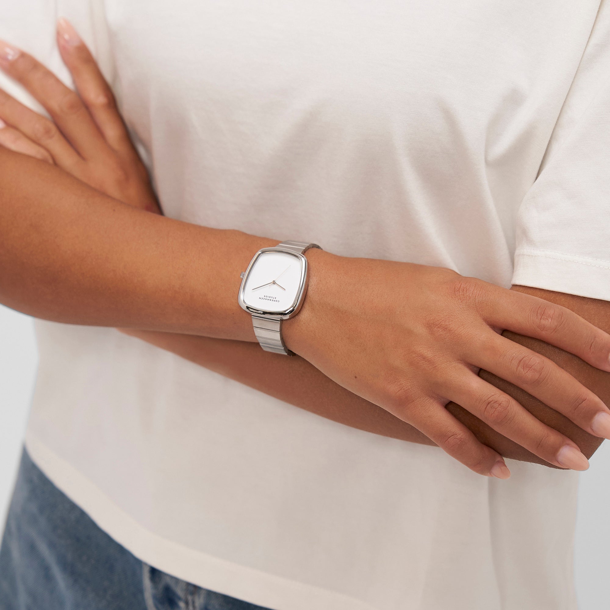Person, die ein weißes Copenhagen Studios T-Shirt und die Copenhagen Studios Armbanduhr - Curved Watch mit über dem Oberkörper verschränkten Armen trägt.