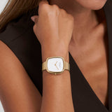 Eine Frau trägt eine Copenhagen Studios Armbanduhr - Curved Watch, eine goldene Armbanduhr mit weißem Ziffernblatt und ohne Zahlen, an ihrem linken Handgelenk.