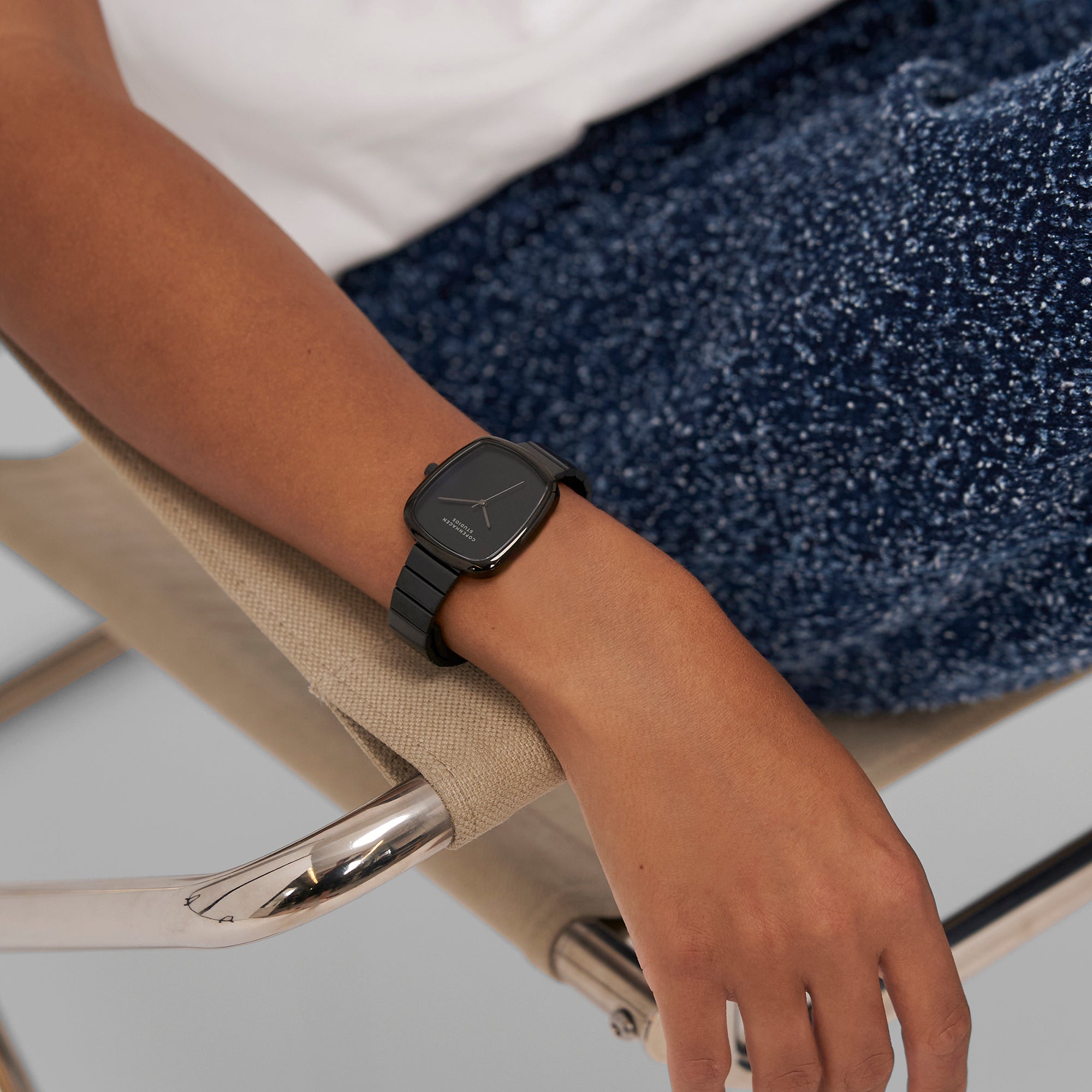Eine Person, die die Copenhagen Studios Curved Watch trägt, sitzt auf einem beigen Stuhl und ist mit einer blauen, strukturierten Hose bekleidet.