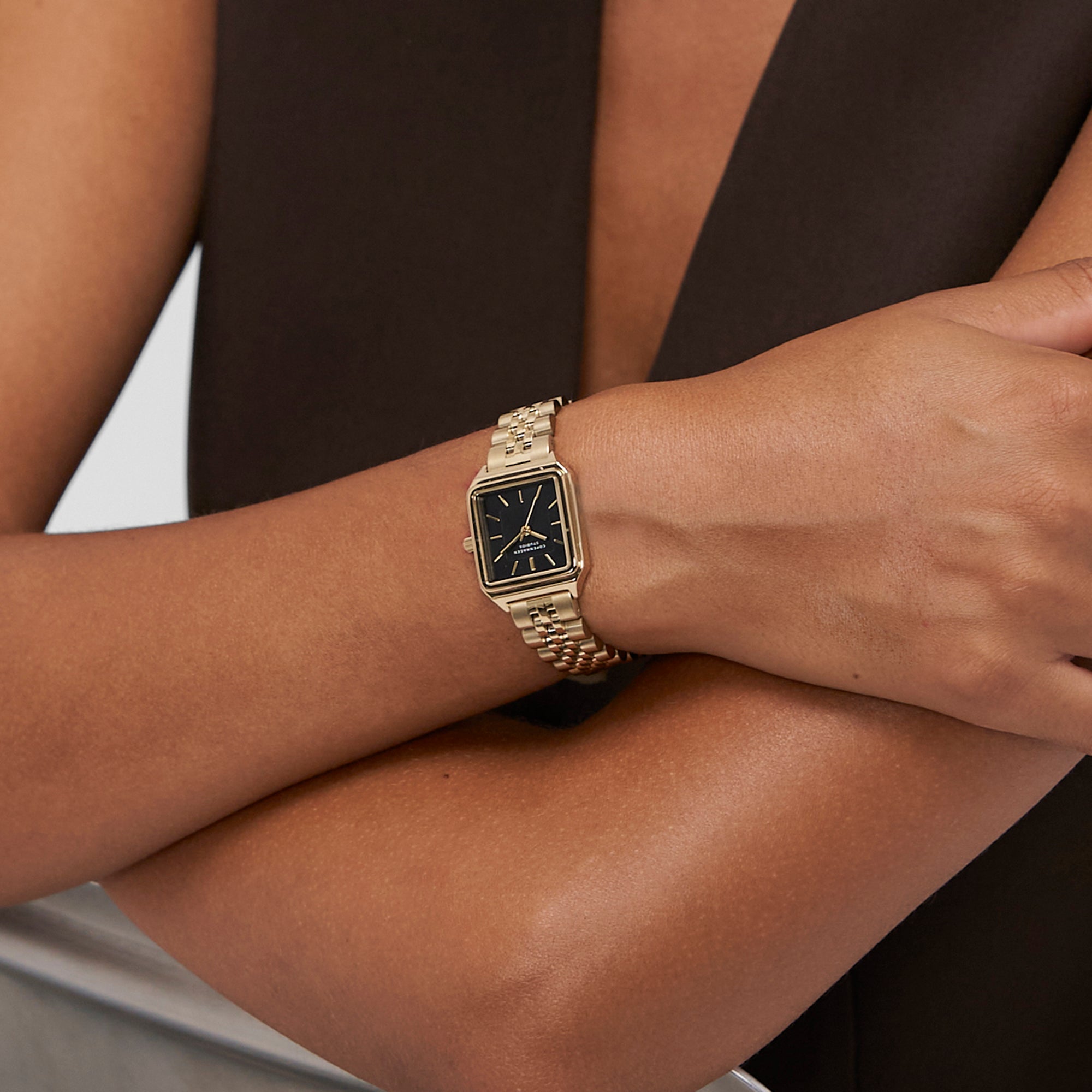 Jemand, der die COPENHAGEN STUDIOS Armbanduhr - Vintage Watch von Copenhagen Studios mit goldenem Band und schwarzem Zifferblatt zu einem ärmellosen, dunklen Top trägt.