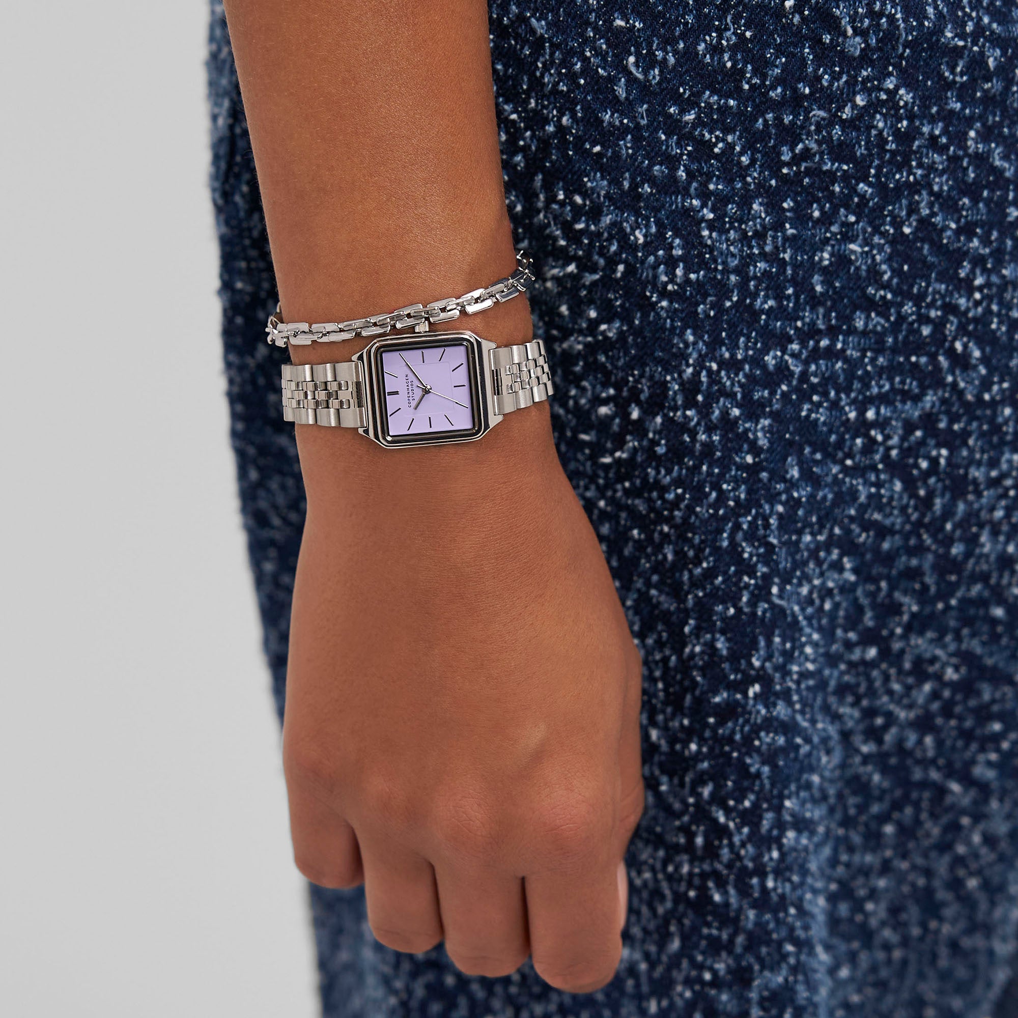 Eine Person trägt eine Copenhagen Studios Armbanduhr - Vintage Watch mit lila Zifferblatt und silbernem Armband, gepaart mit einem blau strukturierten Outfit.