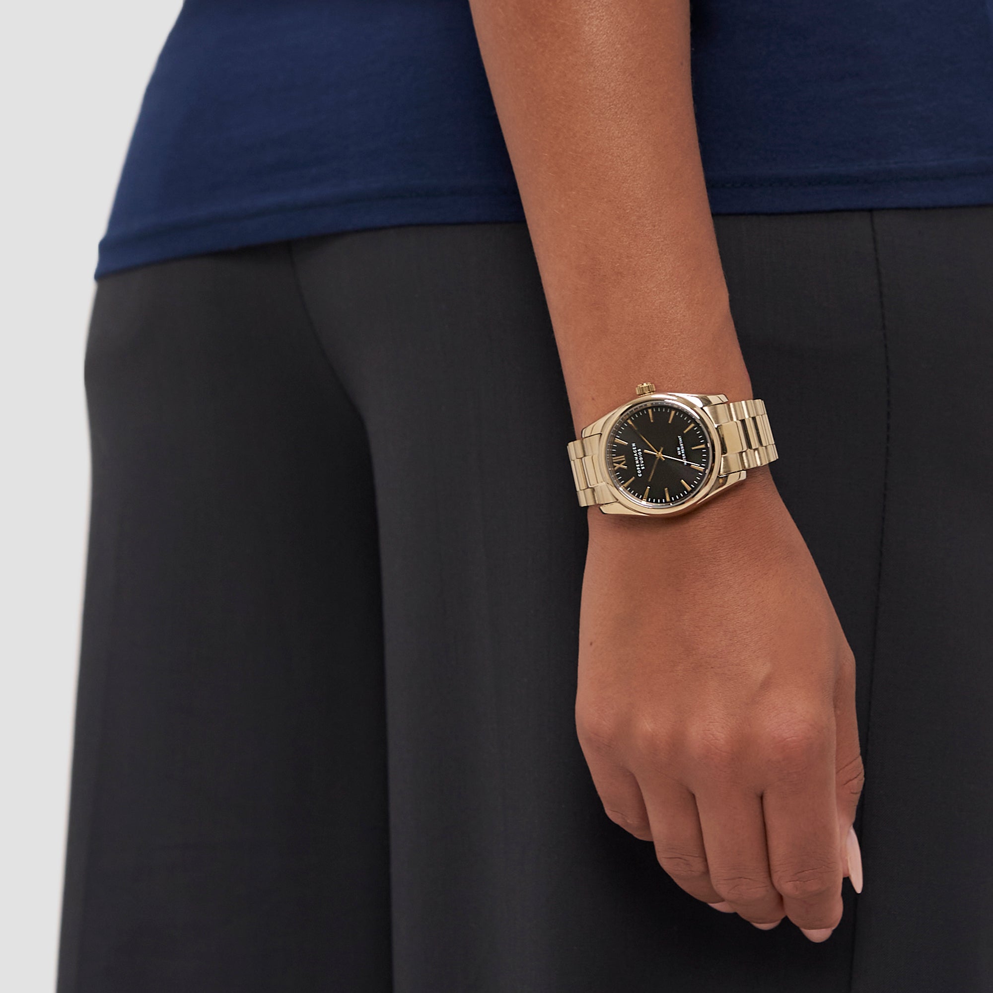 Eine Person trägt die COPENHAGEN STUDIOS Armbanduhr - Round Watch von Copenhagen Studios mit goldenem Gehäuse und schwarzem Ziffernblatt an ihrem linken Handgelenk, gepaart mit einer dunklen Hose und einem marineblauen Top für einen eleganten Look.