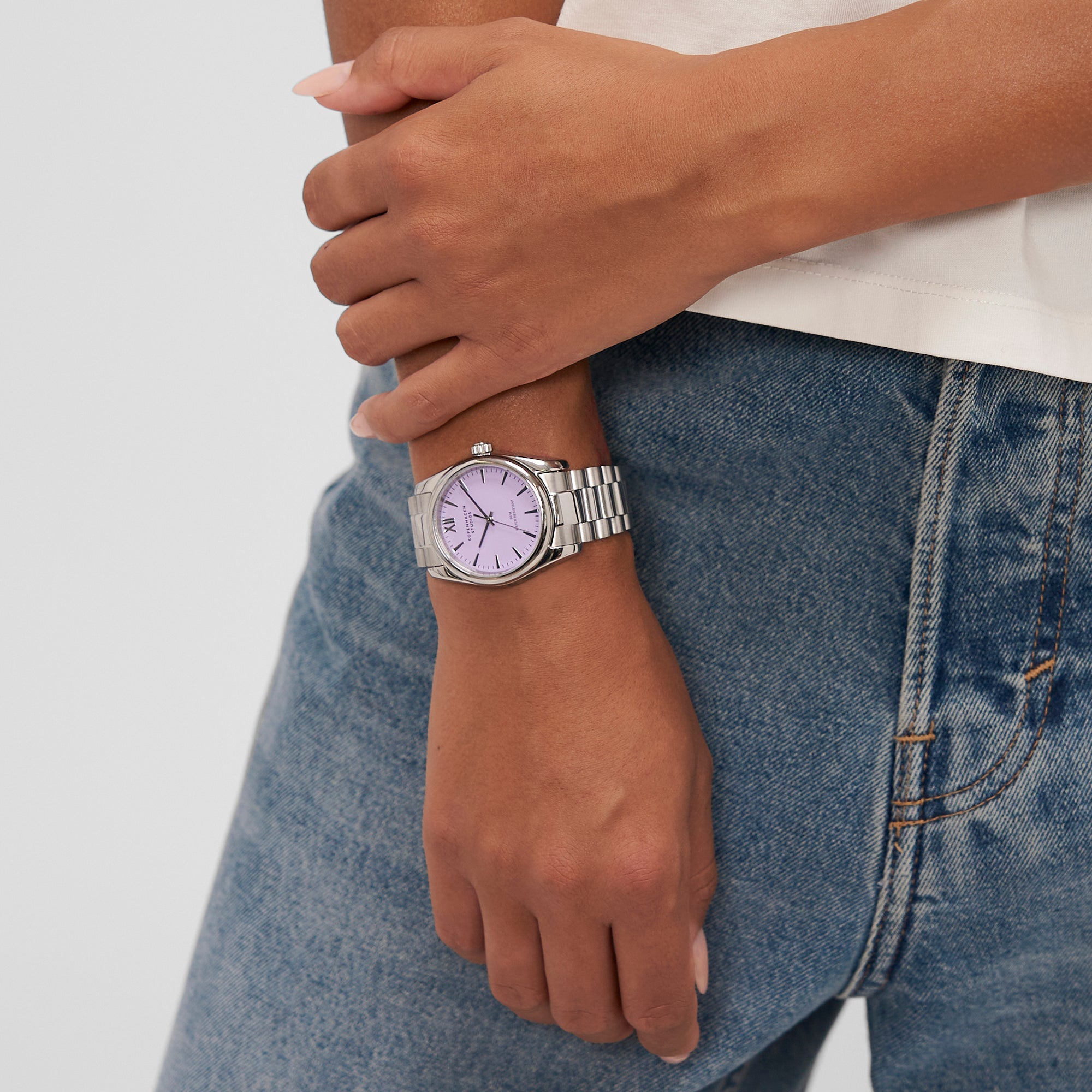 Eine Person trägt die COPENHAGEN STUDIOS Armbanduhr - eine klassische runde Uhr von Copenhagen Studios mit silbernem Band und lavendelfarbenem Zifferblatt, die perfekt zu Blue Jeans und einem weißen Hemd passt.