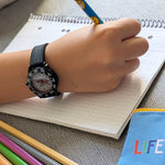 Ein Kind, das die Cool Time Kids Armbanduhr - The Cool Black Racer von Cool Time trägt, schreibt in ein Notizbuch mit einem Bleistift, neben Buntstiften und einem blauen Etui.