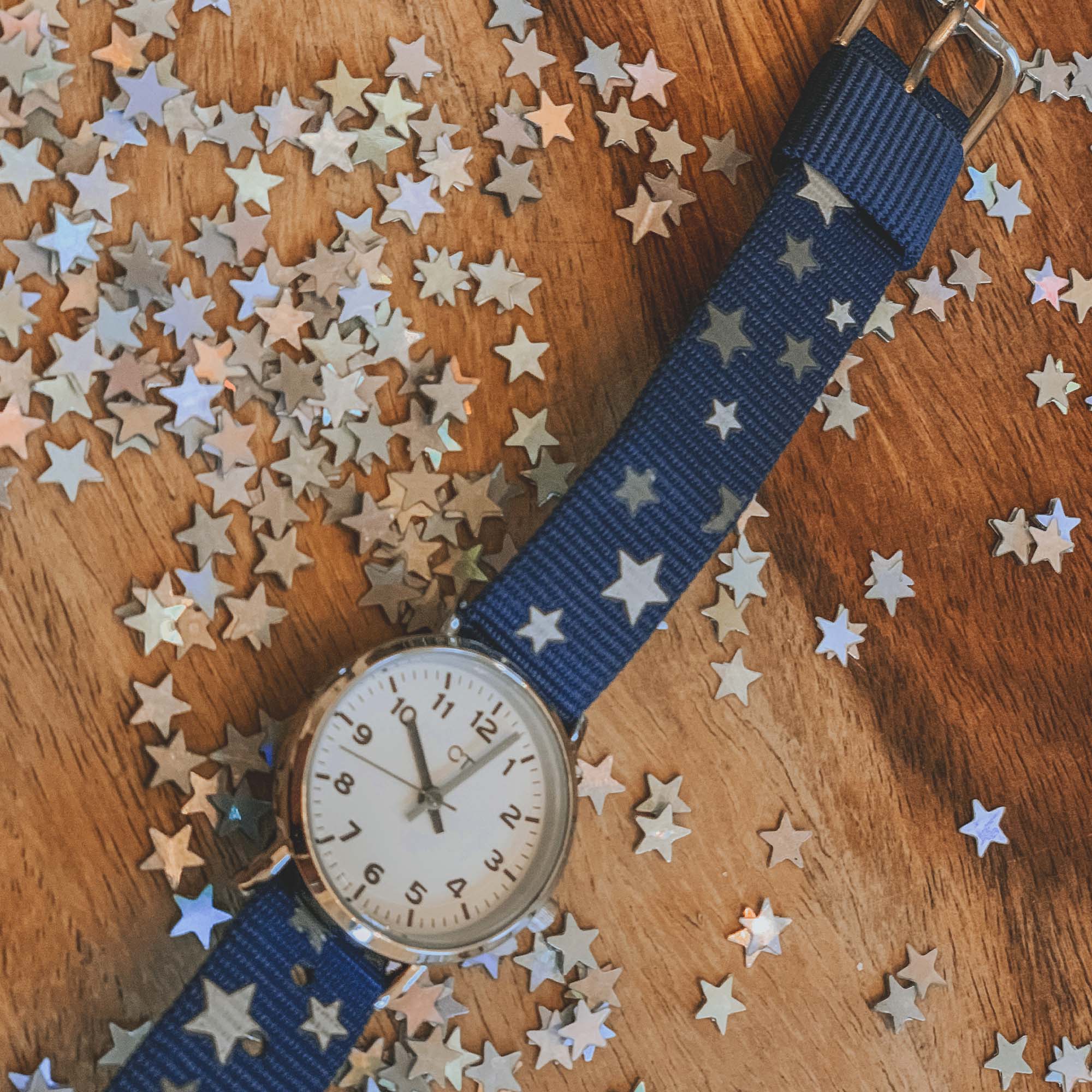 Analoge Uhr mit blauem Armband auf einer hölzernen Oberfläche, umgeben von verstreutem silbernem Sternenkonfetti.