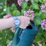 Eine Hand, die die Cool Time Kids Armbanduhr - The Cool Flower Power von Cool Time trägt, hält eine grüne Gießkanne neben rosa Hortensienblüten.